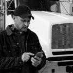 A royalty free image from the trucking and logistics industry of a truck driver standing in front of his vehicle using a smartphone to text or surf the internet.